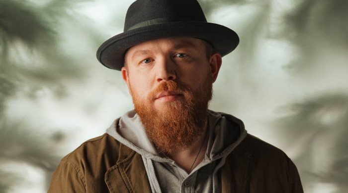 A man with a ginger beard wearing a black hat, grey jumper and green jacket